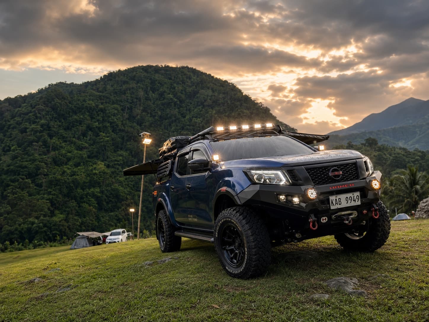 Prototype vehicle with roof rack and bumper lighting on at a hillside campsite at sunset
